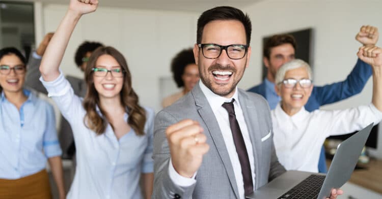 Group of diverse business professionals celebrating success, smiling and raising their fists in excitement, with a man in a gray suit holding a laptop in the foreground.
