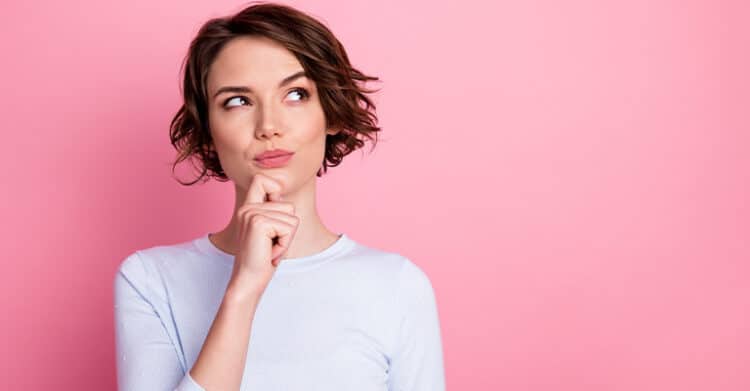 18 Ways to Write Better Blog Posts That Readers Will Finish 6 A young woman with short brown hair stands against a solid pink background, resting her chin on her hand and gazing upward in thought. Her curious expression visually represents brainstorming or planning, aligning with the creative process behind writing better blog posts.