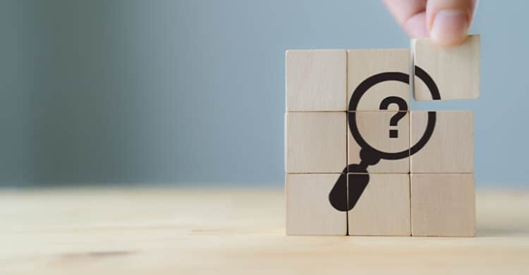 18 Ways to Write Better Blog Posts That Readers Will Finish 11 Wooden blocks arranged in a square display a magnifying glass with a question mark inside, with one block being lifted by a hand. This image symbolizes problem-solving and discovery, aligning with the research and insight needed to create better blog posts that answer readers’ questions.