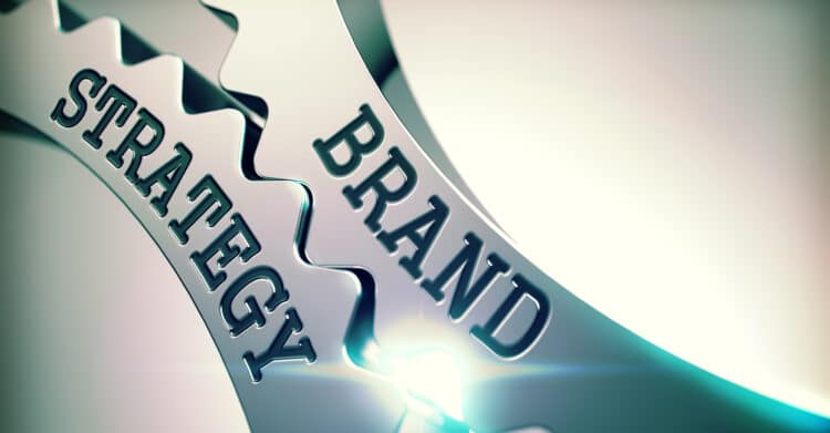 Close up of interlocking metal gears engraved with the words Strategy and Brand, fitting together with a bright highlight at the connection point. The image represents alignment and planning, supporting content about how clear strategy helps Improve Your Brand through cohesive direction and execution.