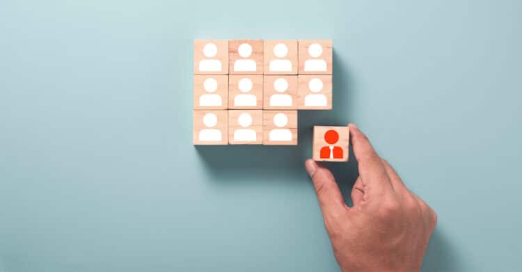 Effective Copywriting: The Core Pillars That Convert 4 Hand placing a wooden block with an orange person icon into a grid of matching blocks on a blue background, suggesting team selection or hiring. The simple visual supports effective copywriting about identifying the right audience or choosing the right person for a role.