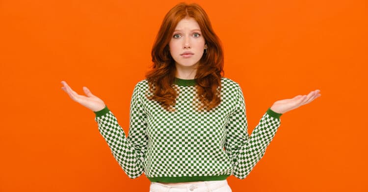 A young woman with long red hair and a green checkered sweater stands against a bold orange background, shrugging with an uncertain expression. Her confused gesture visually represents common frustrations or questions people face when learning SEO copywriting.