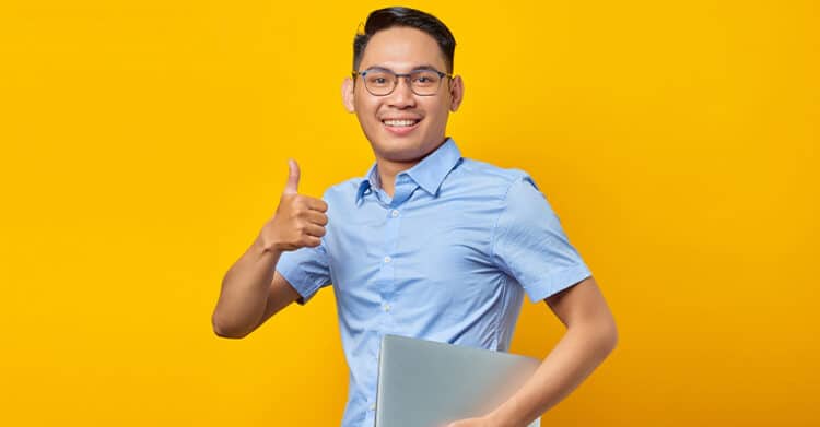 Smiling young man in a light blue shirt and glasses stands against a bright yellow background, holding a closed laptop and giving a thumbs up. The image conveys confidence and satisfaction, often used to represent success or approval—possibly in the context of mastering SEO copywriting or digital work.