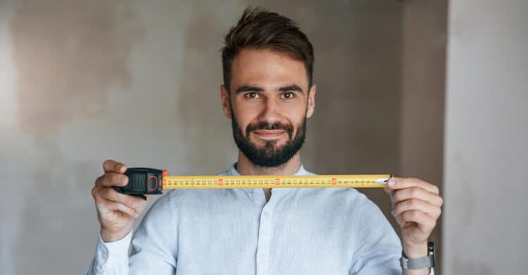How Does Keyword Length Impact SEO Ranking? 3 A man with a neatly groomed beard and short dark hair stands indoors, holding a yellow tape measure stretched out in front of him. He wears a light blue button-up shirt and smiles slightly while looking directly at the camera.