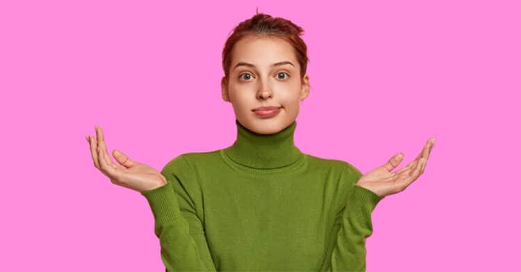 A young woman wearing a green turtleneck sweater stands against a bright pink background with both hands raised and an unsure expression. The image conveys confusion, uncertainty, or indifference in a lighthearted or casual way.
