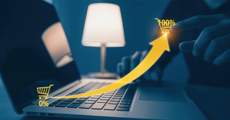 A glowing yellow arrow curves upward from 0% to 100% across a laptop keyboard, ending at a shopping cart icon being tapped by a user's finger. The image symbolizes growth in online sales, conversion rates, or e-commerce performance.