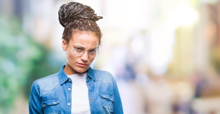 A woman wearing glasses and a denim shirt stands with a serious expression, furrowed brow, and slightly pursed lips, conveying skepticism, frustration, or disapproval. The background is softly blurred, drawing focus to her facial expression.
