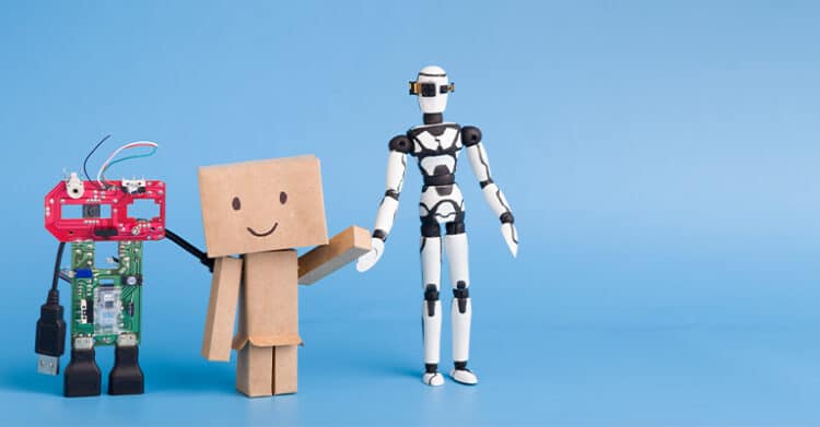Three toy robots stand side by side against a light blue background, each representing a different stage of technological evolution: one made from exposed circuit boards and USB parts, one resembling a simple cardboard box with a smiling face, and one sleek and humanoid in black and white. The image humorously illustrates the progression of robotics and AI design.