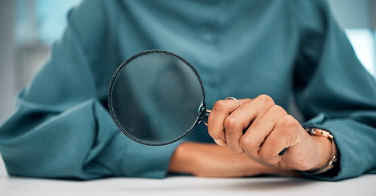 A person in a teal shirt holds a magnifying glass close to the camera, focusing attention on the act of searching, examining, or analyzing. The image symbolizes investigation, research, or scrutiny, commonly associated with SEO, audits, or data review.