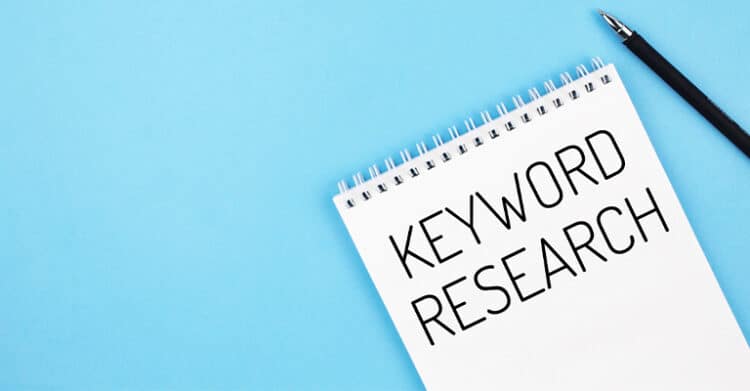 A spiral notebook with the words "KEYWORD RESEARCH" written in bold black letters lies on a light blue background next to a black pen. The image visually represents the concept of SEO planning and digital content strategy.