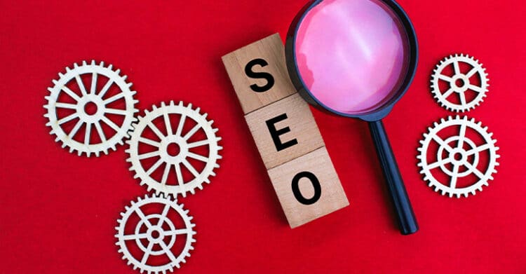 Wooden blocks stacked vertically spell out "SEO," surrounded by light-colored gear cutouts and a pink magnifying glass on a vivid red background. The image symbolizes the technical and analytical components of SEO strategy and optimization.