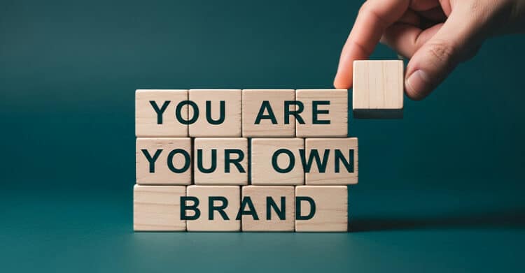 A hand places a wooden block onto a stack of blocks that spell out YOU ARE YOUR OWN BRAND in bold black letters. The teal background and building blocks emphasize personal branding as something you actively create and shape.