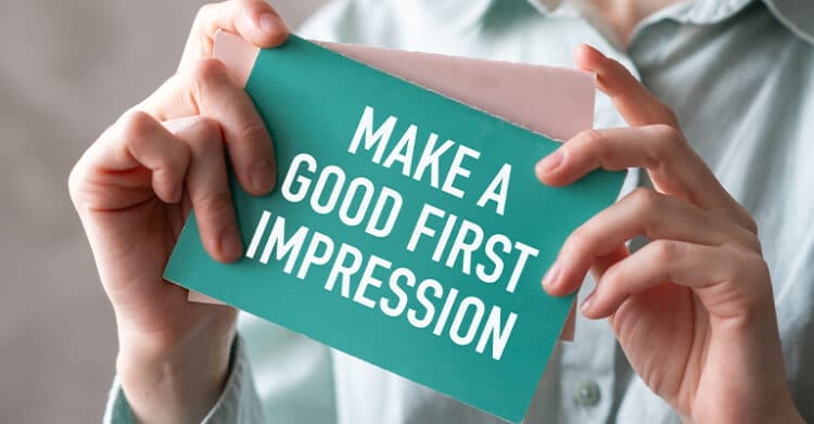 A person holds a teal card that says MAKE A GOOD FIRST IMPRESSION in large white letters. The close up framing focuses on the message as a reminder about presenting yourself well.