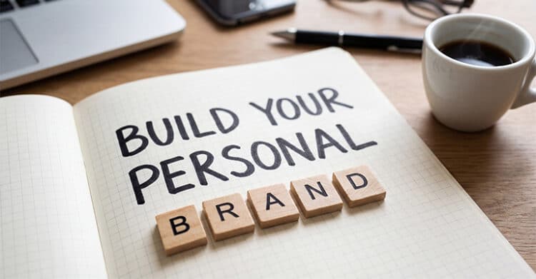 An open notebook on a wooden desk reads BUILD YOUR PERSONAL BRAND with letter tiles spelling BRAND across the page. A coffee cup, pen, and laptop in the background suggest a workspace focused on personal branding work.