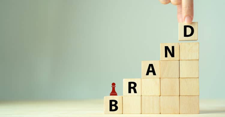 A hand places the final wooden block on a staircase of blocks spelling BRAND, with a small red pawn on the lower step. The rising steps and letters suggest building a brand over time through steady progress and strategy.