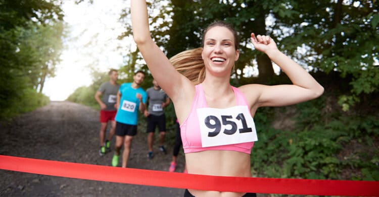 A smiling runner wearing bib number 951 raises her arms as she crosses a red finish line on a wooded trail. Other race participants run behind her, highlighting the moment of completing a race and celebrating the finish.