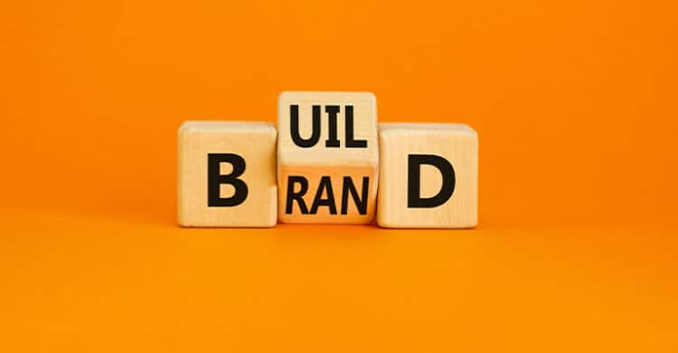 Personal Branding Strategy: Build a Brand That Opens Doors 5 Wooden letter blocks arranged on an orange background spell out the phrase build brand across multiple cubes. The image visually represents branding, brand building, or developing a clear identity in a bold and simple way.