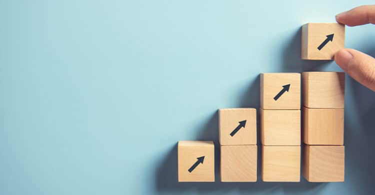 Personal Branding Strategy: Build a Brand That Opens Doors 7 Wooden blocks stacked like a staircase show black arrows pointing upward on each block against a light blue background. A hand places the top block, symbolizing progress, growth, or building success step by step.