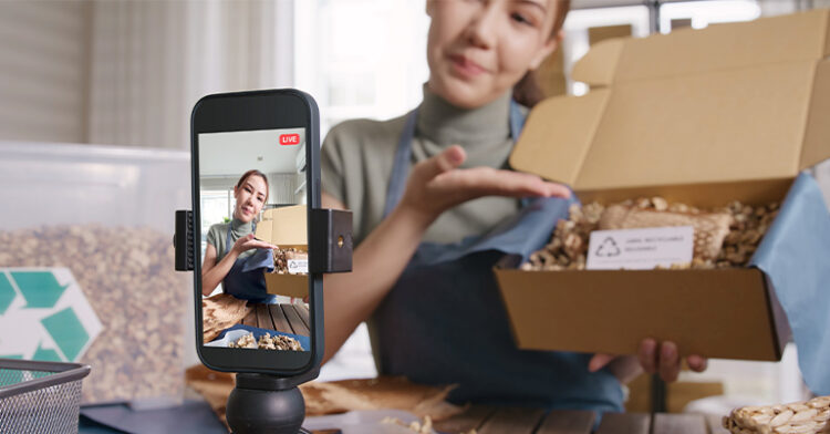 A woman livestreaming product packaging while explaining its eco-friendly features, with her smartphone on a stand.