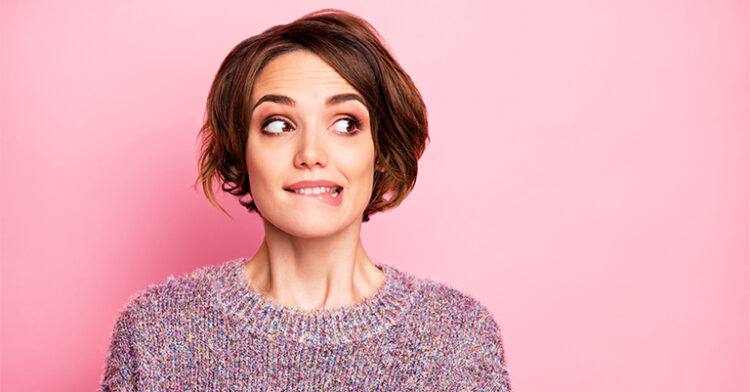 A woman with short hair and a nervous smile bites her lip, looking off to the side against a pink background.