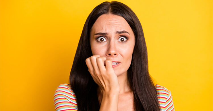 A worried woman biting her nails, with a nervous expression, standing against a bright yellow background.