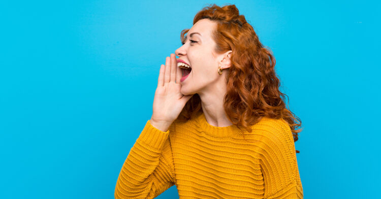 A cheerful red-haired woman in a mustard sweater shouting playfully, standing against a vibrant blue background.