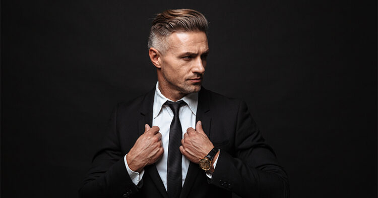 A confident man in a black suit adjusting his tie, exuding professionalism and style against a dark background.