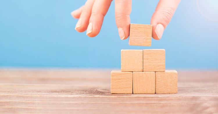 A hand carefully placing a wooden block on top of a small pyramid of stacked wooden cubes.