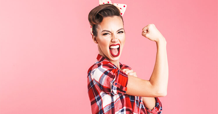 Woman flexing her bicep with a fierce expression, wearing a red plaid shirt and retro polka-dot headband, standing in front of a pink backdrop.