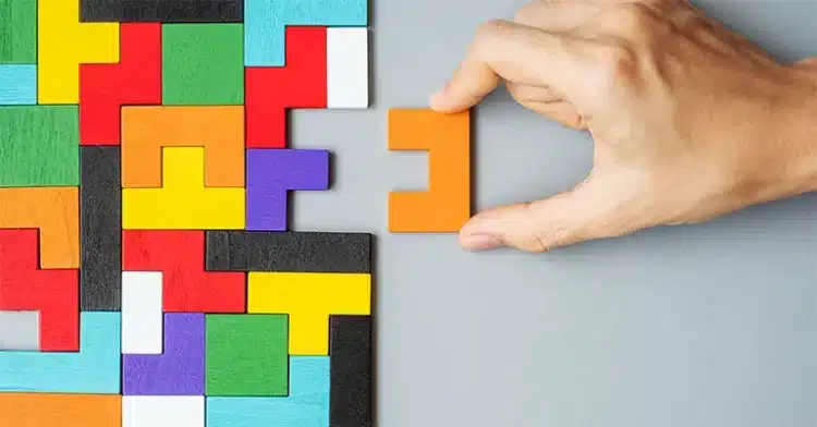 Hand placing an orange puzzle piece into a nearly completed multicolored puzzle on a gray surface.