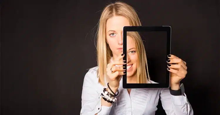 How to Manage Your Online Identity: The Ultimate Guide 3 Woman holding a tablet that shows her smiling face while her real expression behind it appears serious.