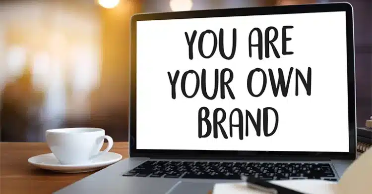 Laptop screen displaying the phrase "YOU ARE YOUR OWN BRAND" next to a coffee cup on a wooden table.