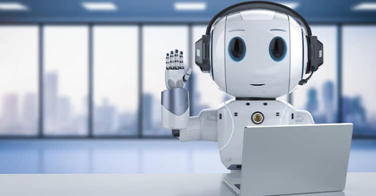 A humanoid robot wearing headphones sits at a desk with a laptop, raising its hand as if greeting someone during a virtual conversation.