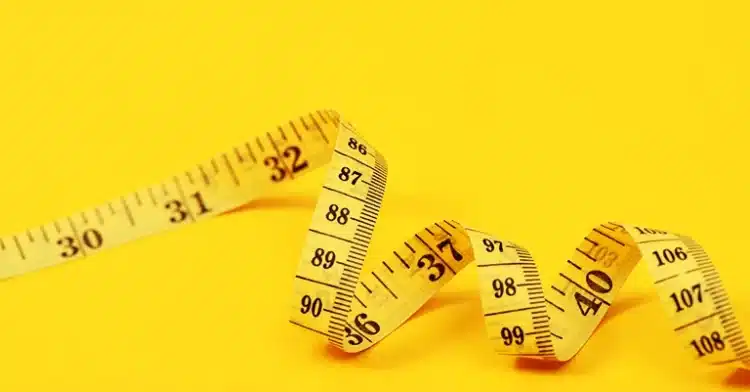 A close-up, angled shot of a yellow measuring tape lying on a bright yellow background. The tape is slightly curled and shows various numerical markings in black, including 30, 31, 32, 36, 37, 40, 86, 87, 88, 89, 90, 97, 98, 99, 103, 106, 107, and 108. The image highlights the texture and detail of the measuring tape against the solid, vibrant background.