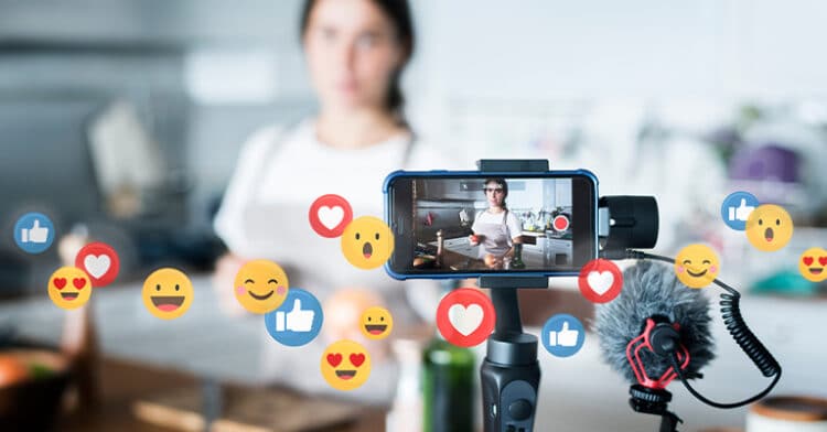 A woman livestreaming in her kitchen with a smartphone setup, surrounded by floating social media reaction icons.