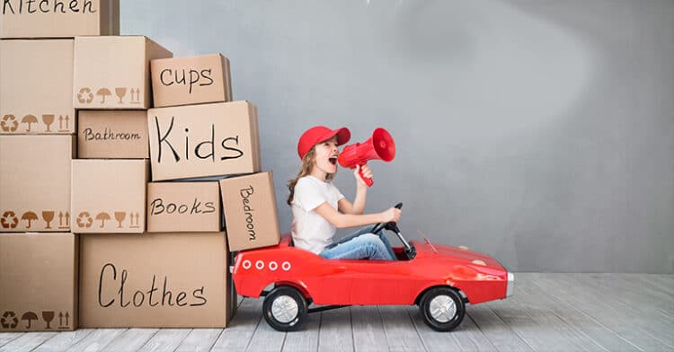 Child in red toy car shouting into a megaphone beside moving boxes labeled “Kids,” “Clothes,” “Books,” and more.
