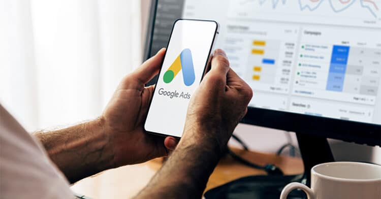 A person holds a smartphone displaying the Google Ads logo in front of a computer screen showing ad performance data and campaign metrics.