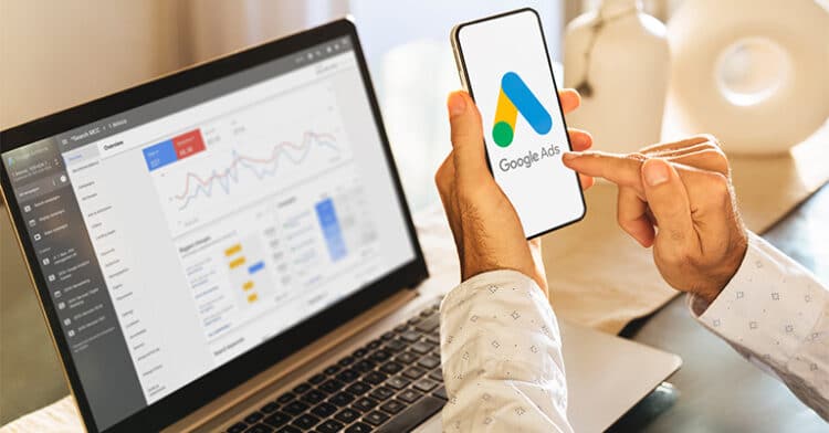 Marketer holding a smartphone with the Google Ads logo displayed while reviewing campaign analytics on a laptop screen during a digital strategy session.