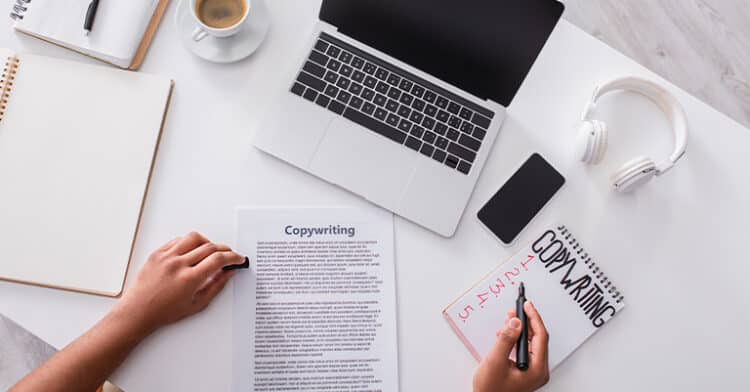 Person working on copywriting at a white desk with a laptop, headphones, coffee, and notes, suggesting content creation and digital marketing tasks.