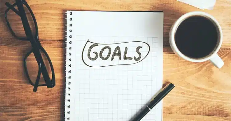 How to Use Social Media Influencers to Improve Your Marketing 6 Notebook with the word “GOALS” written on graph paper, next to a pen, eyeglasses, and a cup of coffee on a wooden desk.