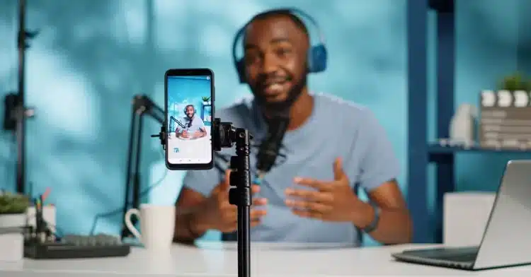 Man wearing headphones speaks into a microphone while recording a video with a smartphone on a tripod.