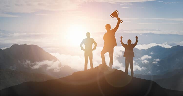 Silhouetted business team on a mountaintop at sunrise, with one person holding a trophy in victory.