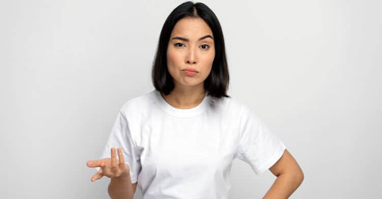 Woman in a plain white t-shirt standing against a neutral background with a confused and slightly skeptical expression, raising one hand in a questioning gesture. Her body language suggests uncertainty or disbelief.