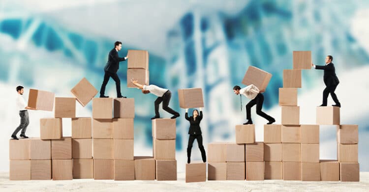 Group of businesspeople in formal attire working together to stack and balance large wooden cubes, symbolizing teamwork and collaboration. The background is blurred with blue and white tones, emphasizing the focus on the coordinated effort in the foreground.