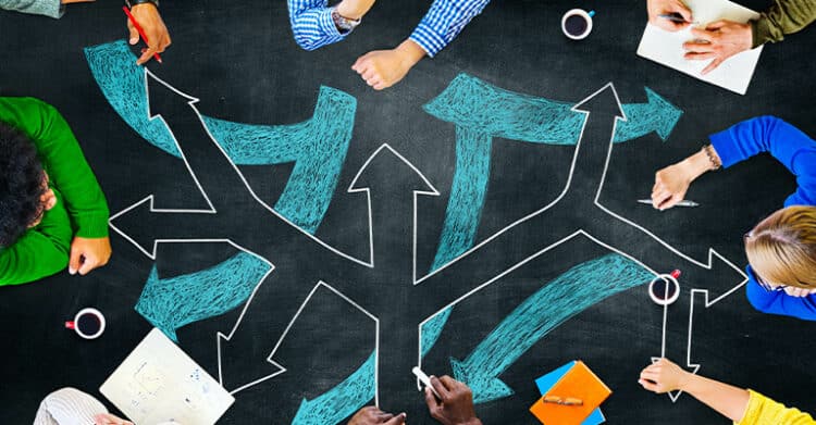Overhead view of a diverse group of people sitting around a black table with white and teal arrows drawn in multiple directions, symbolizing decision-making or choosing a path. Hands are shown holding pens, papers, and coffee cups, indicating a collaborative brainstorming or planning session.