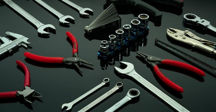 Flat lay of assorted hand tools on a glossy black surface, including wrenches, pliers with red handles, socket set, caliper, and a tape measure. The tools are neatly arranged, emphasizing precision, variety, and readiness for mechanical or technical tasks.