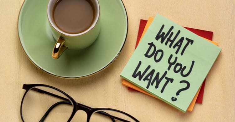 A flat lay shows a coffee cup on a saucer beside eyeglasses and stacked sticky notes on a desk. The top note reads what do you want, suggesting reflection, goal setting, or clarifying priorities in a calm workspace setting.
