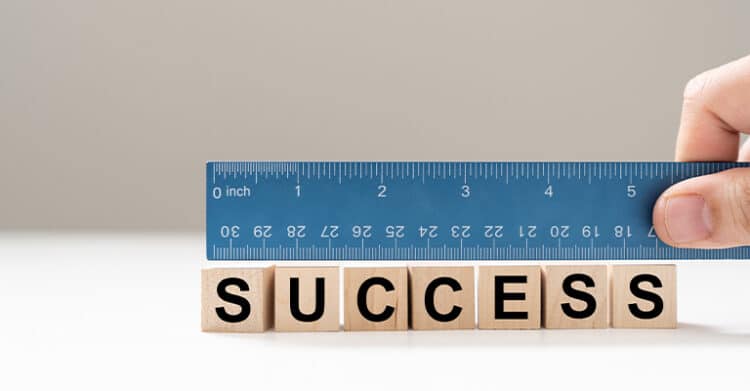 Wooden letter blocks spelling the word success sit on a white surface while a hand holds a blue ruler above them. The image suggests measuring success, evaluating results, or defining progress using clear metrics.