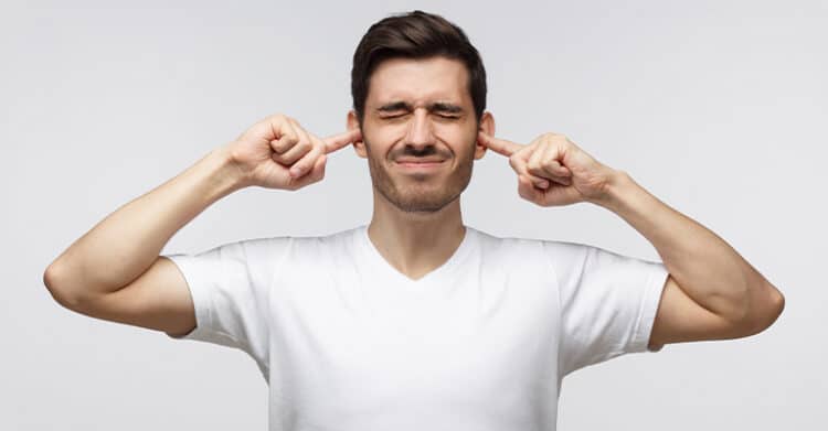 A man wearing a white t shirt presses his fingers into his ears with his eyes closed against a neutral background. The image conveys blocking out noise, annoyance, or avoiding unwanted information or distractions.