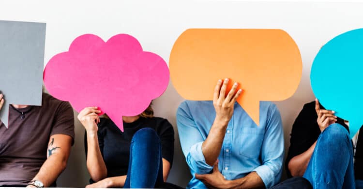 A group of people sit side by side holding large colorful speech bubble cutouts in front of their faces. The image represents communication conversation feedback or diverse voices being shared without focusing on individual identities.